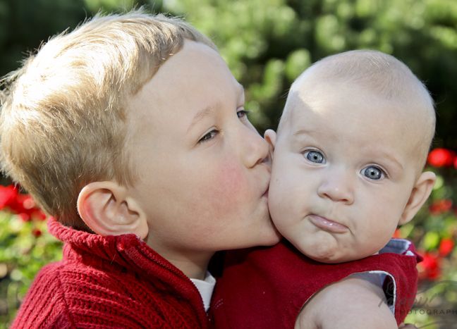 A young child affectionately kisses a baby on the cheek, with a background of vibrant red flowers