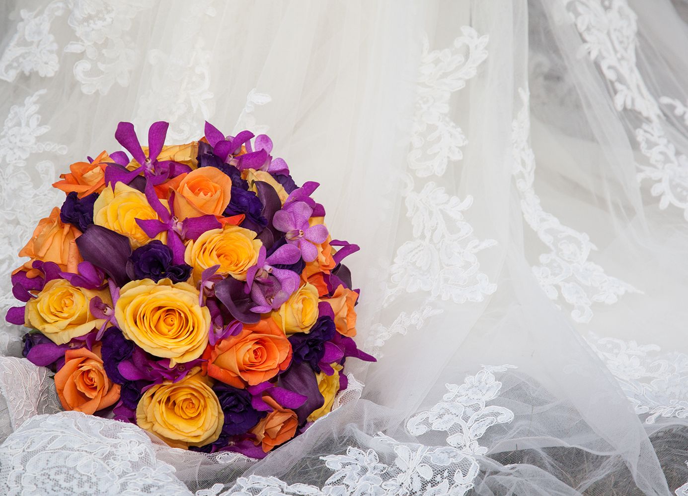 A colorful bouquet of flowers with orange and yellow roses, purple orchids set against a veil with intricate lace patterns.
