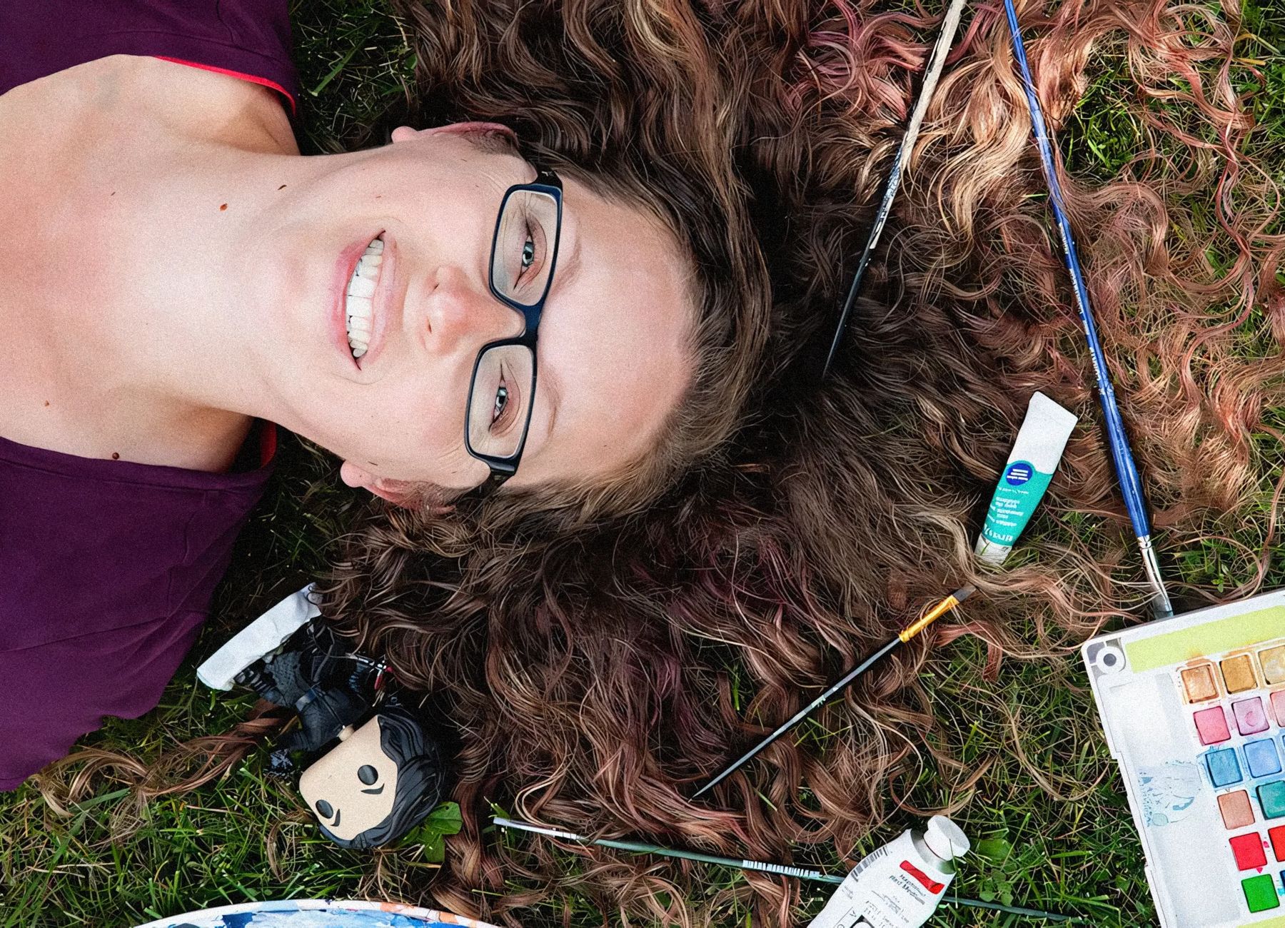 A person lying down on the grass, surrounded by art supplies including a doll, paint tubes, and brushes.
