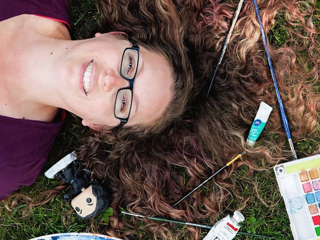 A person lying down on the grass, surrounded by art supplies including a doll, paint tubes, and brushes.